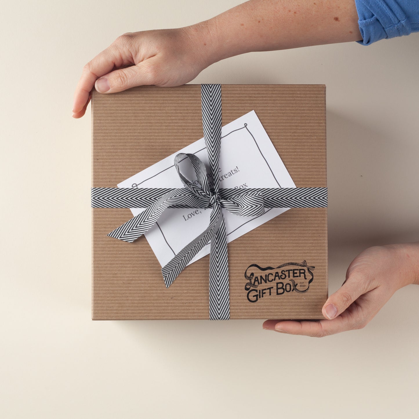 Hands presenting a kraft Lancaster Gift Box with herringbone ribbon and gift note–ships gift-ready nationwide.