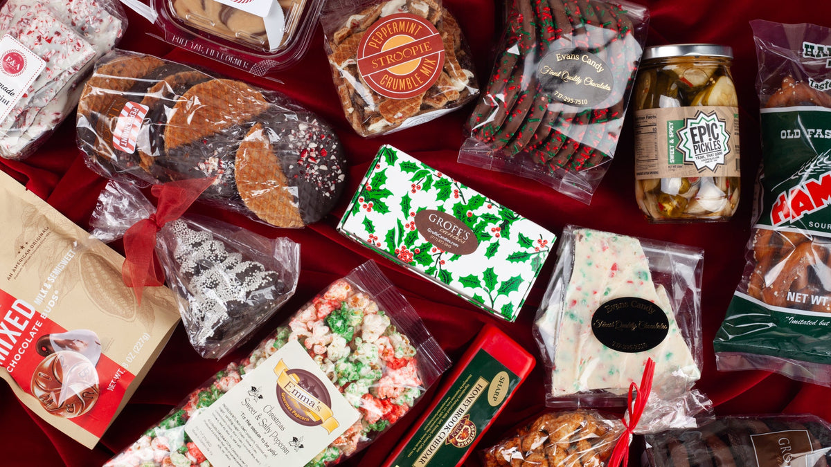 Lancaster Gift Box holiday collection spread on red velvet, featuring peppermint stroopwafels, Groff's Candies holly box, Emma's Christmas popcorn, peppermint bark, Hammond's pretzels, and seasonal Pennsylvania treats.