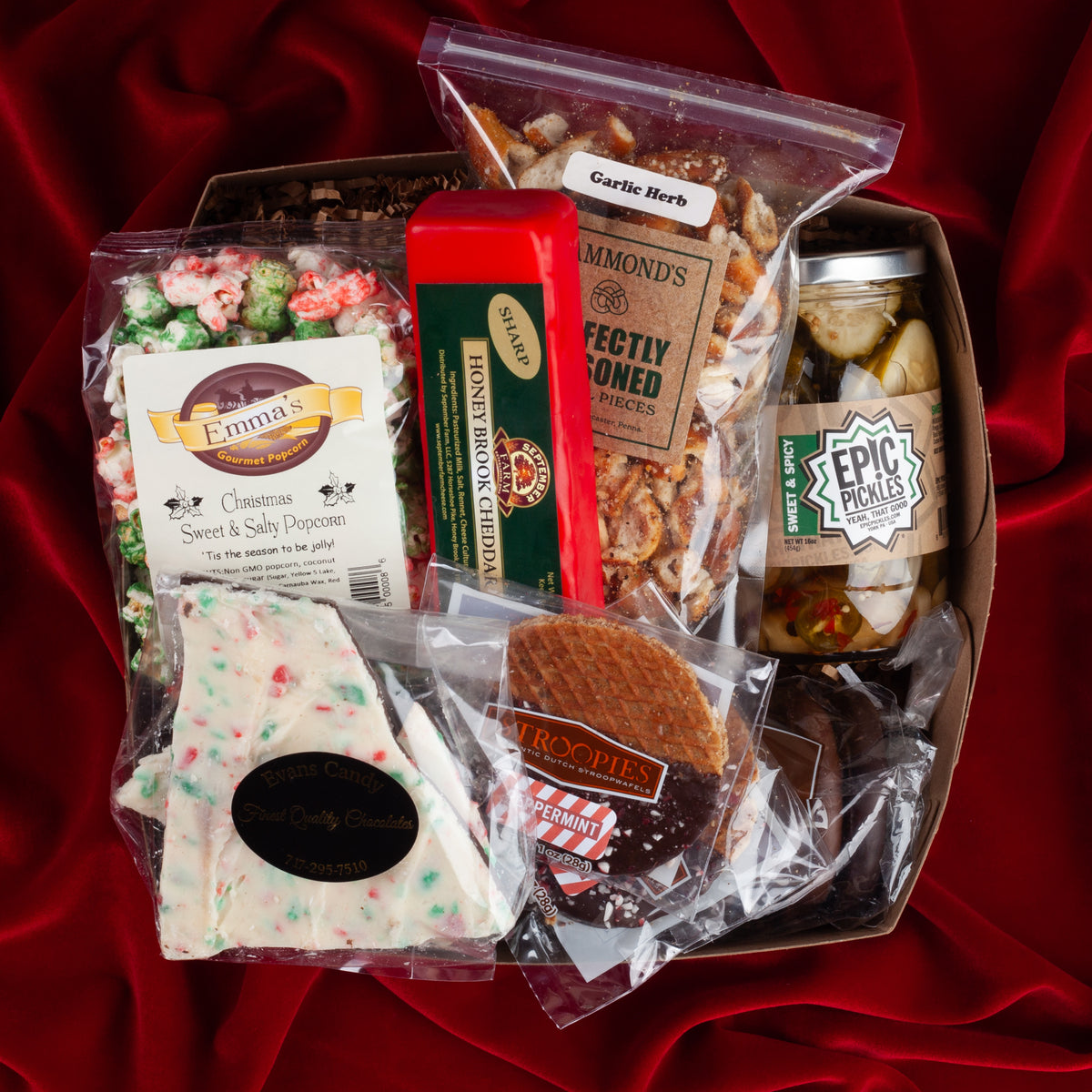 Holiday Cheer gift box on a red velvet background, filled with seasonal popcorn, peppermint bark, festive pretzels, peppermint stroopwafels, honey brook cheddar in red wax, and sweet and spicy pickles from Pennsylvania makers.