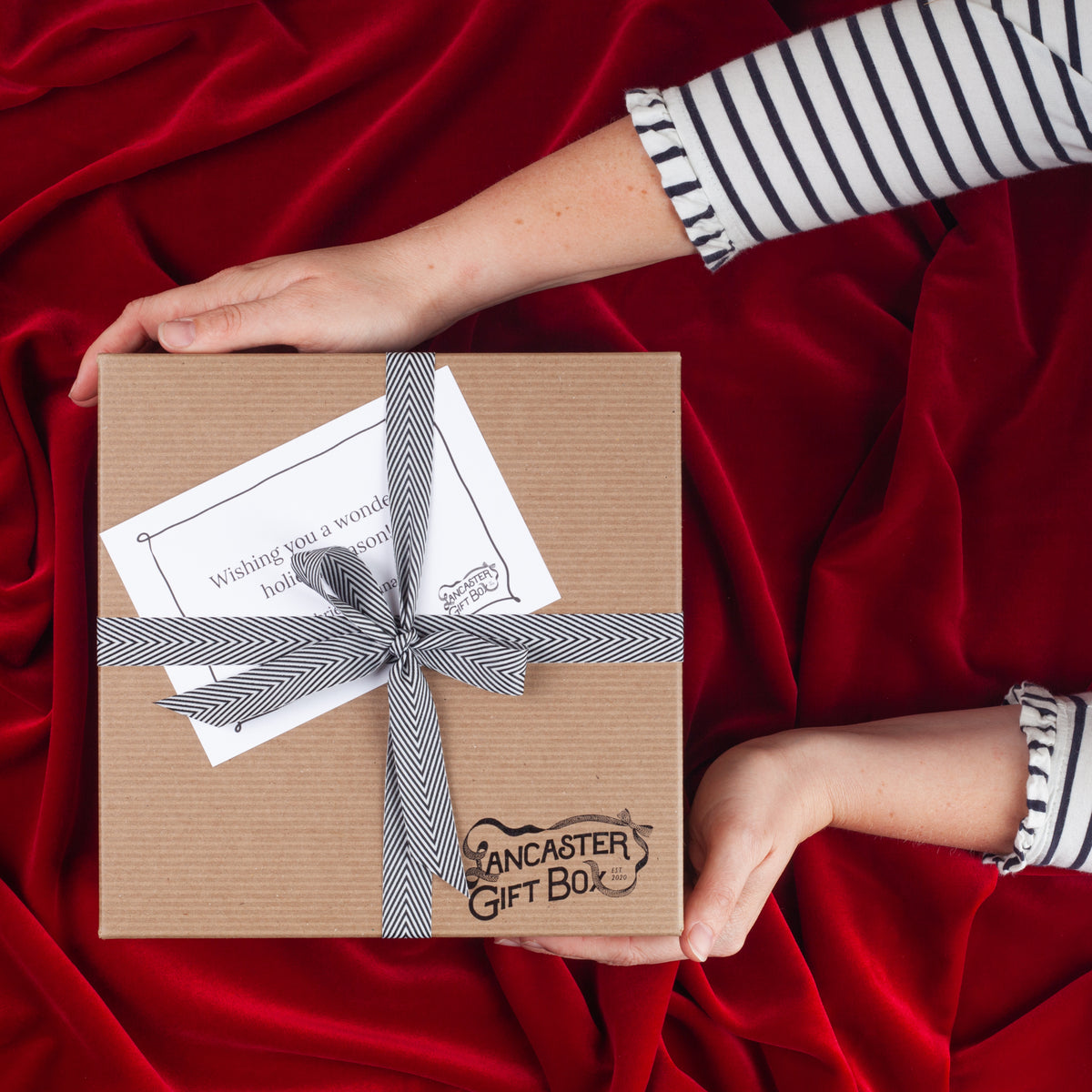 Lancaster Gift Box holiday gift box with black and white herringbone ribbon and handwritten holiday gift card presented on red velvet background.