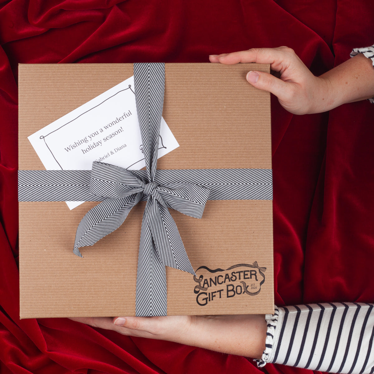 Hands holding a Lancaster Gift Box holiday gift box tied with a black and white herringbone ribbon and personalized gift card on red velvet.