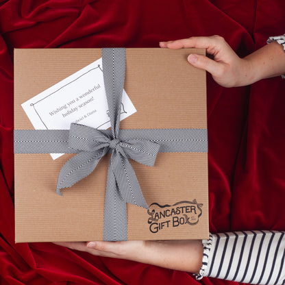 Hands holding a Lancaster Gift Box holiday gift box tied with a black and white herringbone ribbon and personalized gift card on red velvet.