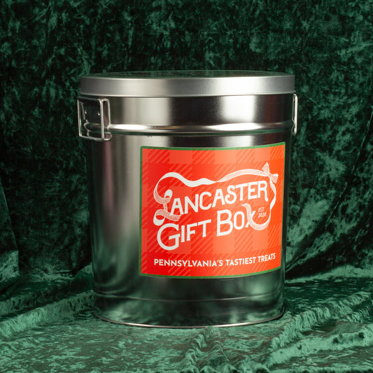Holiday Snack Tin on a green velvet background, a keepsake tin filled with eleven Pennsylvania small-batch holiday treats including peppermint bark, Wilbur Buds, cheddar, pretzels, and festive chocolates.