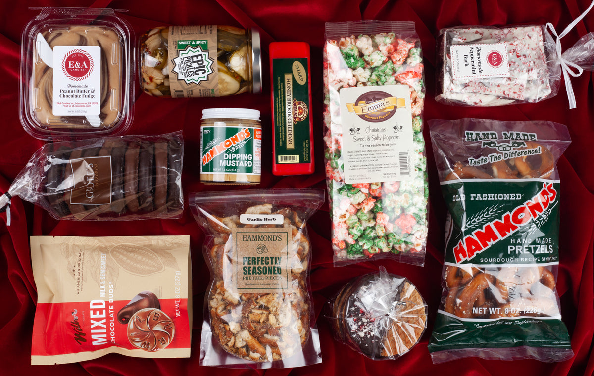 Holiday Snack Tin contents spread on red velvet, featuring Hammond's pretzels, Wilbur Buds, Epic Pickles, September Farm cheddar, Emma's Christmas popcorn, peppermint bark, and fudge from Lancaster, PA.